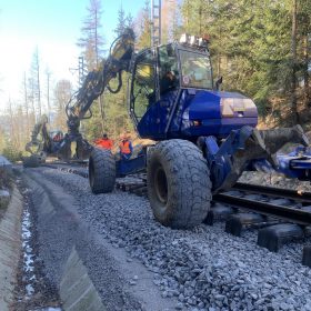 Montáž kolejového roštu pomocí dvou kráčejících bagrů Menzi-Muck se speciálními drapáky pro uchycení kolejnice
