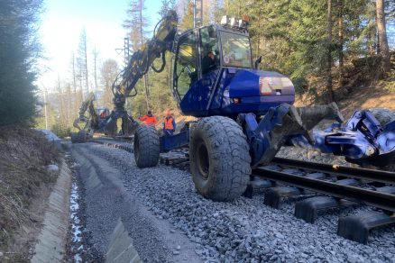 Montáž kolejového roštu pomocí dvou kráčejících bagrů Menzi-Muck se speciálními drapáky pro uchycení kolejnice