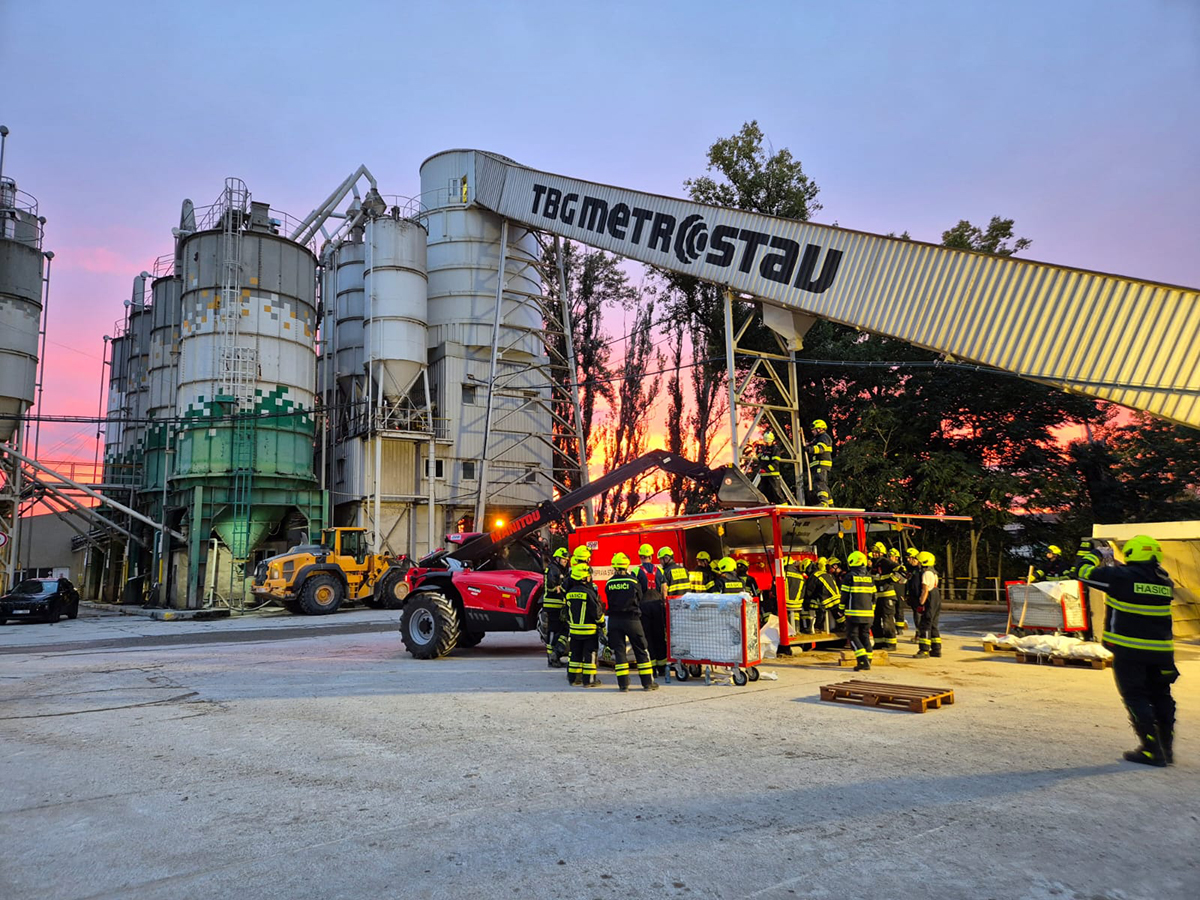 Plnění pytlů pískem v betonárně při západu slunce. Velké nádrže na beton jsou dominantním prvkem prostoru, zatímco hasiči v reflexních uniformách provádějí zásah vedle červeného zásahového vozu. Scéna má dramatickou atmosféru s barevnými odlesky zapadajícího slunce.