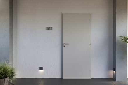 Minimalistické vchodové dvere v svetlosivej farbe na chodbe moderného bytového domu s betónovými prvkami
