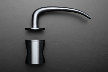 Detailní pohled na moderní designovou kliku z broušené nerezové oceli na tmavém pozadí.