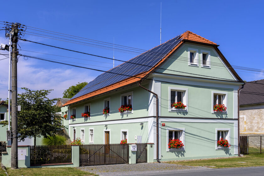 Tradiční rodinný dům s moderní fotovoltaikou na střeše a květinami v oknech.