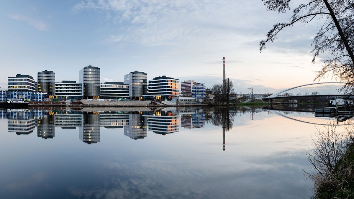 Moderní administrativní budovy u vodní hladiny s odrazem a lávkou v pozadí