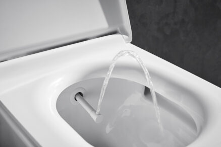 Detail sprchovací toalety bez elektřiny s vysunutou tryskou a proudem vody při oplachu, minimalistické bílé provedení.