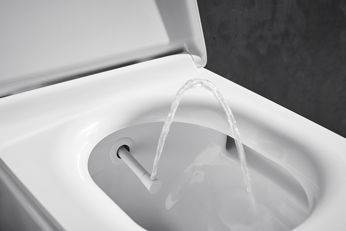 Detail sprchovací toalety bez elektřiny s vysunutou tryskou a proudem vody při oplachu, minimalistické bílé provedení.