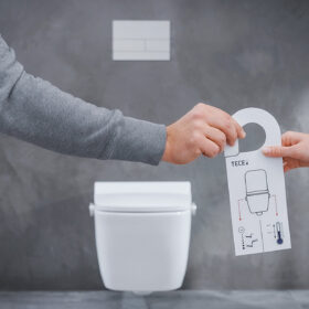 Dvě ruce drží ovládací prvek sprchovací toalety bez elektřiny před závěsnou toaletou v moderní koupelně.