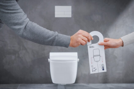 Dvě ruce drží ovládací prvek sprchovací toalety bez elektřiny před závěsnou toaletou v moderní koupelně.