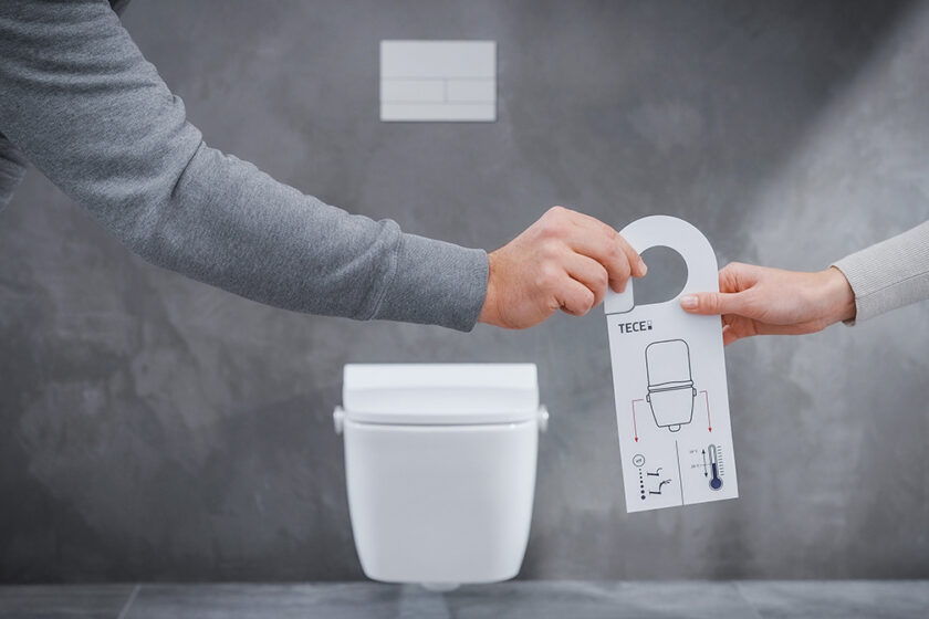 Dvě ruce drží ovládací prvek sprchovací toalety bez elektřiny před závěsnou toaletou v moderní koupelně.