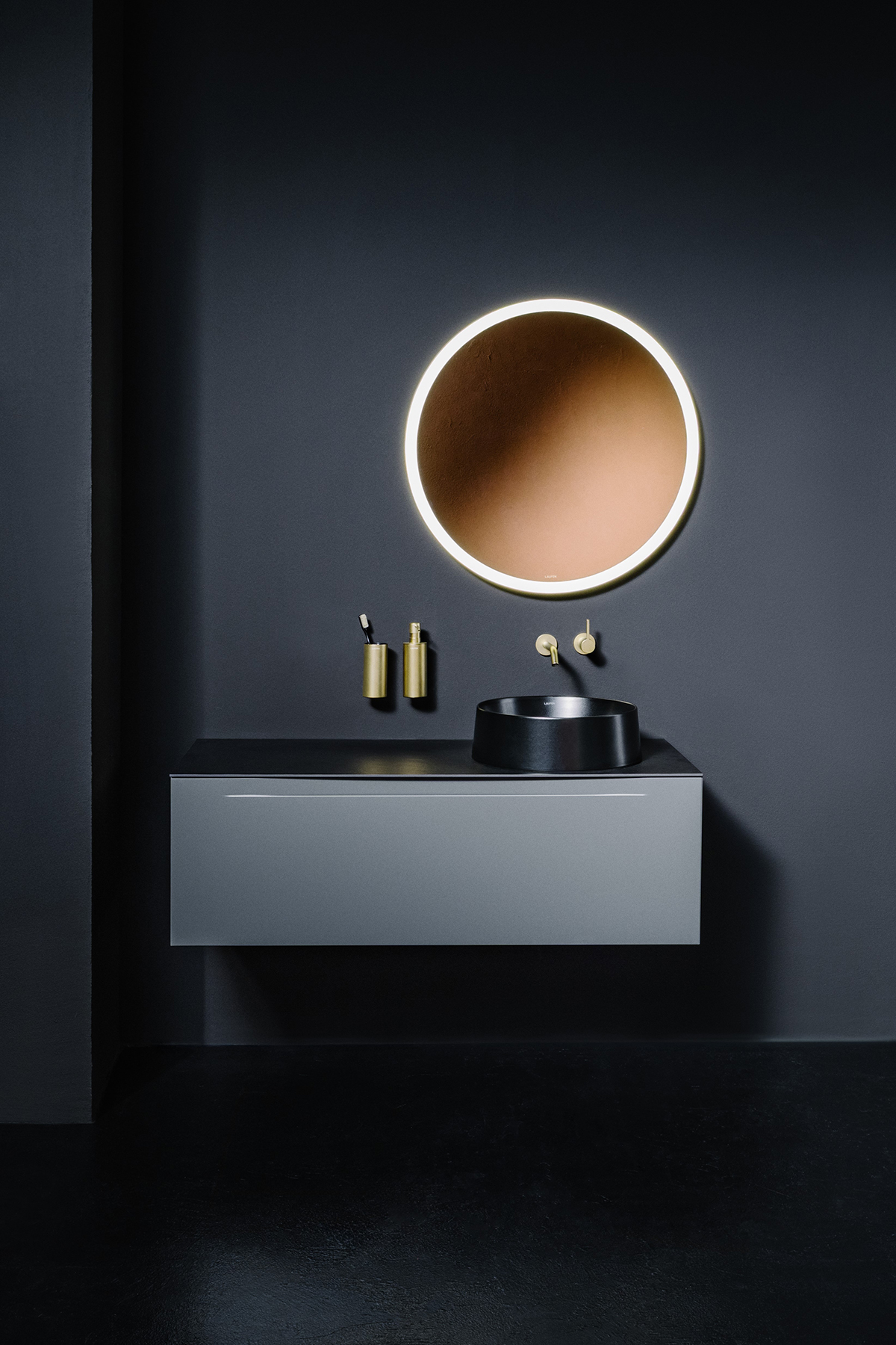Koupelnový interiér s černým umyvadlem, podsvíceným kulatým zrcadlem a mosaznými doplňky v elegantním minimalistickém stylu.