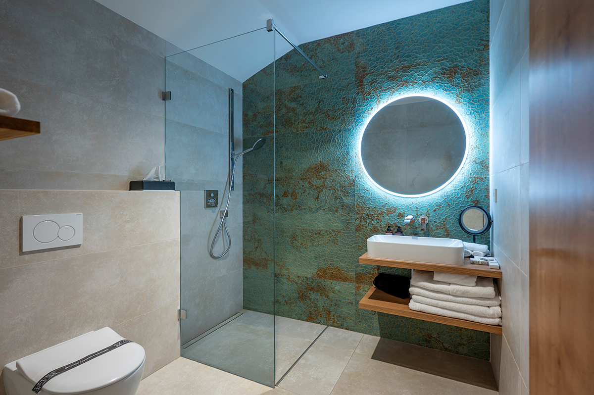 Moderní koupelna hotelu Bronzo se skleněným sprchovým koutem a kulatým zrcadlem